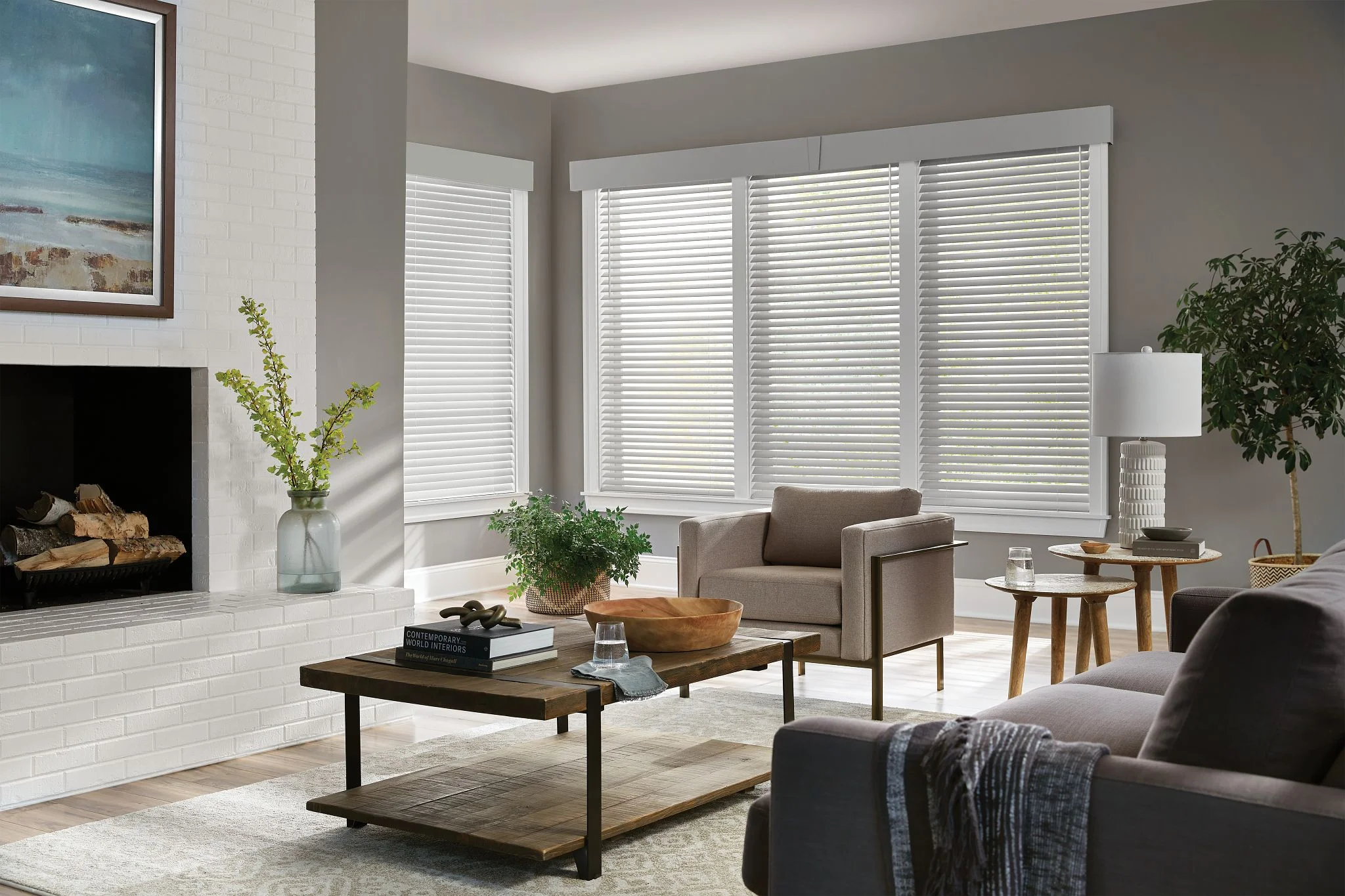 A modern living room featuring a cozy grey sofa, armchair, coffee table, and a brick fireplace with bright natural light filtering through blinds with Shutters & Blinds